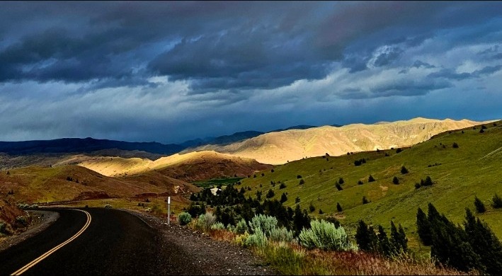 Scenic road curving through sunlit John Day country beneath storm clouds
